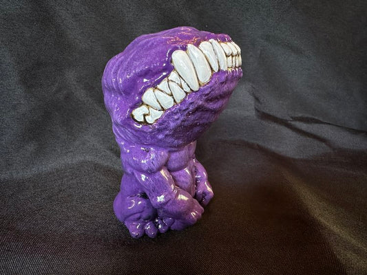 A small figurine. This creature, though humanoid, has no eyes, and its unnaturally large head is defined only by a large, closed mouth with teeth bared. It sits on its hind legs, docile, its head tilted upwards. This version of the doll is a warm purple.
