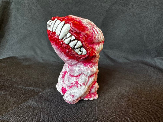A small figurine. This creature, though humanoid, has no eyes, and its unnaturally large head is defined only by a large, closed mouth with teeth bared. It sits on its hind legs, docile, its head tilted upwards. This version of the doll is a pale pink, with vibrant red joints.