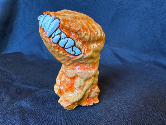 A small figurine. This creature, though humanoid, has no eyes, and its unnaturally large head is defined only by a large, closed mouth with teeth bared. It sits on its hind legs, docile, its head tilted upwards. This version of the doll is a bright orange.
