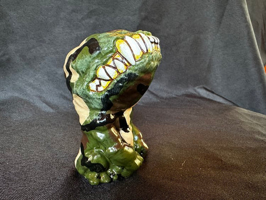 A small figurine. This creature, though humanoid, has no eyes, and its unnaturally large head is defined only by a large, closed mouth with teeth bared. It sits on its hind legs, docile, its head tilted upwards. This version of the doll is green, blank, beige, and brown, creating a camoflauge effect.
