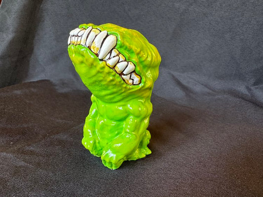 A small figurine. This creature, though humanoid, has no eyes, and its unnaturally large head is defined only by a large, closed mouth with teeth bared. It sits on its hind legs, docile, its head tilted upwards. This version of the doll is a lime green.
