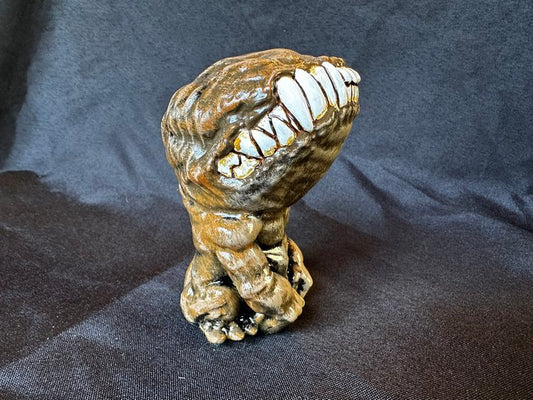 A small figurine. This creature, though humanoid, has no eyes, and its unnaturally large head is defined only by a large, closed mouth with teeth bared. It sits on its hind legs, docile, its head tilted upwards. This version of the doll is a cold brown.