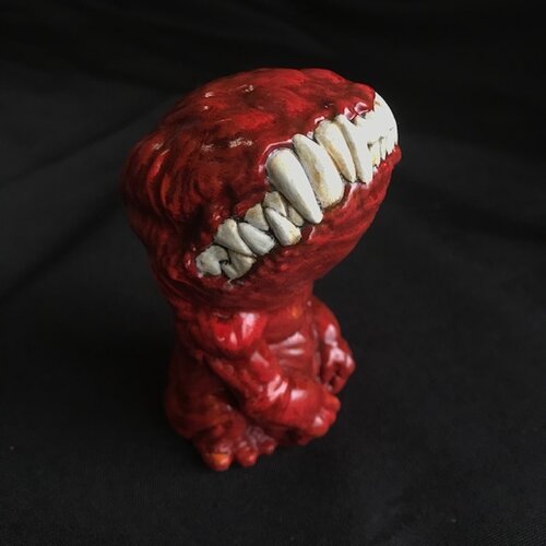 A small figurine. This creature, though humanoid, has no eyes, and its unnaturally large head is defined only by a large, closed mouth with teeth bared. It sits on its hind legs, docile, its head tilted upwards. This version of the doll is a dark red..