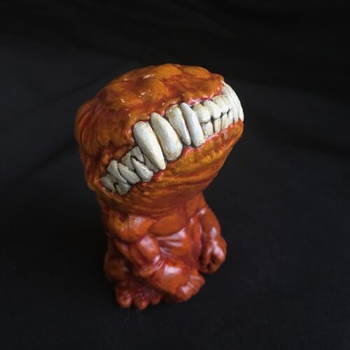 A small figurine. This creature, though humanoid, has no eyes, and its unnaturally large head is defined only by a large, closed mouth with teeth bared. It sits on its hind legs, docile, its head tilted upwards. This version of the doll is a dirty orange.