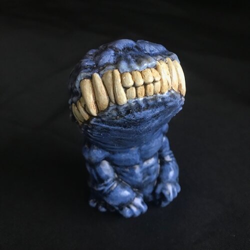 A small figurine. This creature, though humanoid, has no eyes, and its unnaturally large head is defined only by a large, closed mouth with teeth bared. It sits on its hind legs, docile, its head tilted upwards. This version of the doll is a dark blue.